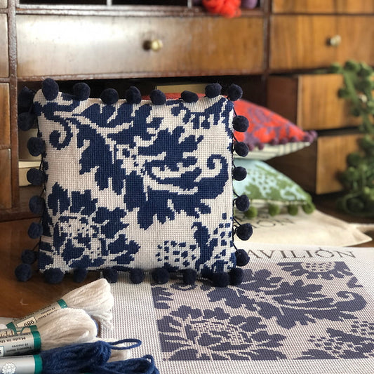 Tapestry cushion with blue floral pattern and pom-poms on a table with needlepoint supplies.